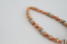 A warm-toned beaded necklace with pops of green and rhinestones, featuring a timeless freshwater pearl bead in the center handmade by artisans overcoming trauma & injustice in Charlotte, NC. 